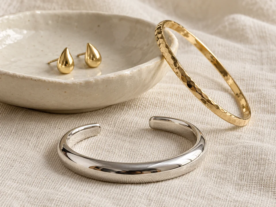 Gold bangle, silver open cuff and teardrop earrings on warm ivory linen and ceramic